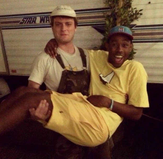 Two men pose playfully together, with the man in the yellow shirt and blue cap carrying the other man (wearing overalls and a "VICK PRESS" cap) in his arms, in front of a trailer displaying a "STAR WAR..." logo.
