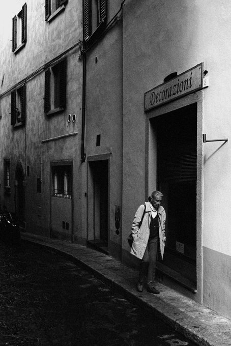 A woman in a trench coat stands in a doorway beneath a sign reading "Decorazioni" on a narrow Italian street, capturing the quiet, intimate atmosphere of old-world European architecture that evokes themes of heritage and cultural lineage. The aged stucco walls, shuttered windows, and worn cobblestone alleyway visually reflect the photographer's exploration of familial roots and homeland, grounding the image in a sense of place and memory.
