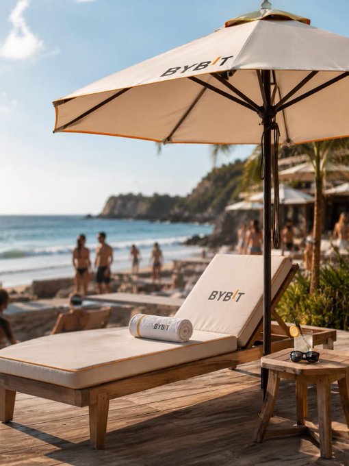 A beachside chaise lounge and umbrella branded with "BYBIT" sit along a sunny tropical shoreline, with people enjoying the sand and ocean in the background. The branded setup aligns with the post's "GM Legends - Go out and touch grass" message, showcasing the crypto exchange's promotional presence in a real-world leisure setting.