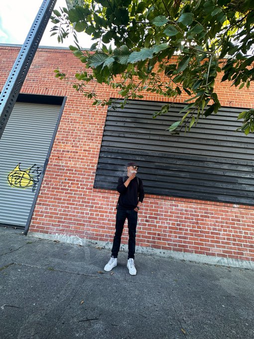 A person dressed in black stands on a sidewalk in front of a red brick building with black paneled windows and a partially visible metal roll-up door featuring a graffiti cat.