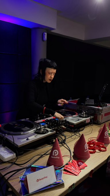 A female DJ performs at a table with two turntables and a mixer, surrounded by 'RedDoPlay' branded party hats and noisemakers.