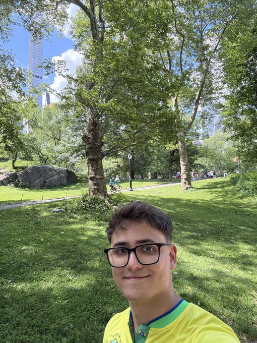 The image features a young man wearing glasses and a yellow shirt who appears to be taking a selfie with his phone. The background of the photo shows a park setting, with trees and grass surrounding him. There are also several other people present in the scene, indicating that this is likely a popular location for outdoor activities or gatherings.
