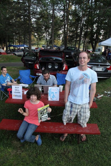 A man and a young girl pose at an outdoor "Buy Sell Learn Bitcoin" booth, with the girl proudly holding a Bitcoin promotional flyer. The scene captures an early grassroots effort to spread awareness and education about cryptocurrency at a time when Bitcoin was still trading around just $5, long before its mainstream recognition.