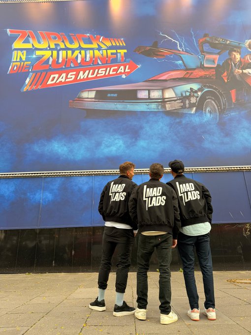 Three people wearing matching black bomber jackets with 'LMAD LADS' printed on the back stand with their backs to the camera, looking at a large poster for 'Zurück in die Zukunft – Das Musical' (Back to the Future The Musical) featuring the DeLorean time machine.