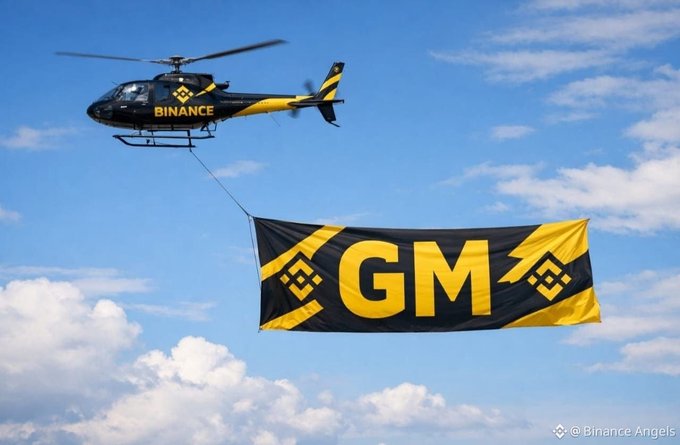 A black and yellow helicopter bearing "BINANCE" branding flies through a partly cloudy sky, towing a large banner displaying "GM" alongside the Binance logo. The image playfully visualizes the crypto community's popular "GM" (good morning) greeting as an aerial announcement, tying the cheerful message to Binance's branding.