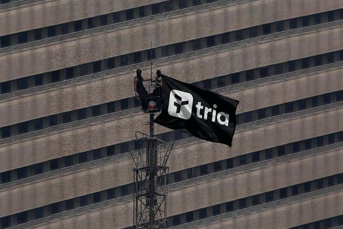 Two workers in a crane basket attaching a large black "tria" branded flag to a pole against the backdrop of a tall building facade.