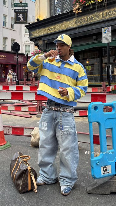 A man wearing a yellow cap, blue and yellow striped rugby shirt, baggy jeans with the number '03', and blue rhinestone shoes eats a slice of pizza on a London street near the Golden Lion pub, with a Louis Vuitton duffel bag beside him. Watermark: @taymoesofficial.