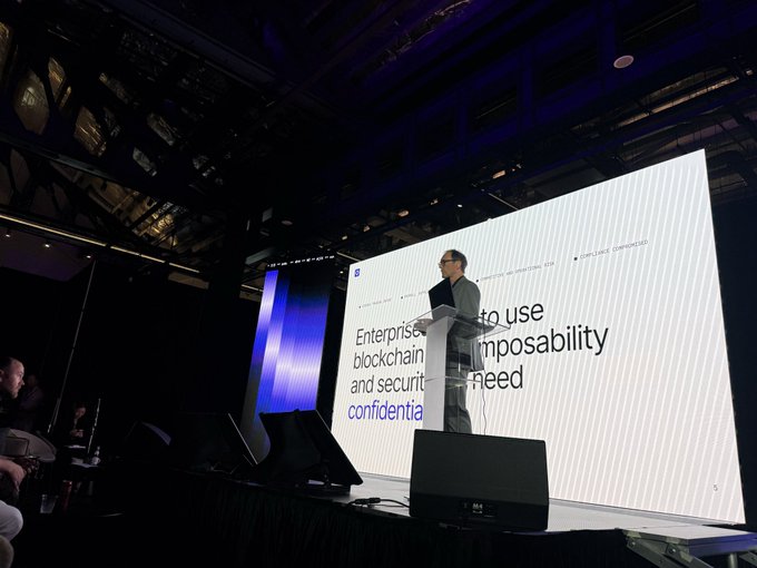 A man stands at a podium on a stage delivering a presentation, with a large screen behind him displaying the text 'Enterprise... to use blockchain... impossibility and secuir... need...' along with a 'POWERED BY CONFIDEO' logo.