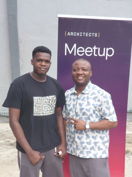 Two young men posing together in front of a purple banner reading '{ARCHITECTS} Meetup'; the man on the left wears a black 'LET IT BE' graphic t-shirt, while the man on the right wears a heart-patterned short-sleeve shirt and makes a peace sign.