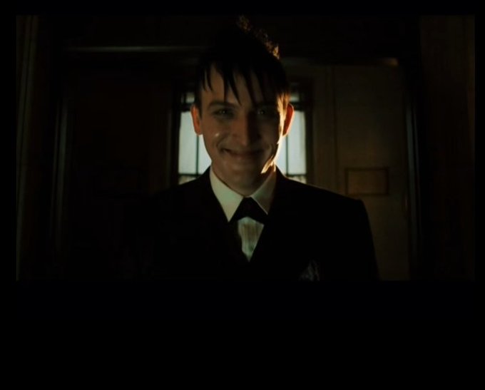 A man in a dark suit and tie smiles menacingly in a dimly lit hallway, appearing to be a still from a film.