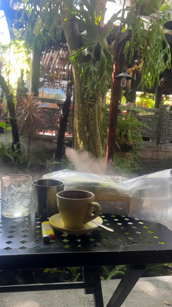 The image features an outdoor patio with a black metal table holding two cups of coffee - one yellow cup and one black cup. A silver spoon rests next to each cup, suggesting that someone has recently enjoyed their beverages. The scene also includes a potted plant in the background, adding a touch of greenery to the setting. The image is related to a post on social media where people are encouraged to share their thoughts about the world and its problems.