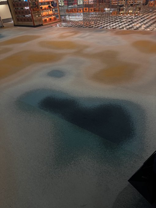 An artistic floor installation with swirling patterns of orange, white, and teal blue resembling an aerial view of sand and water, with a checkered tiled area and display shelves visible in the background.