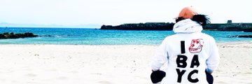 A person sitting on a beach wearing a white hoodie that reads 'I ❤️ BAYC' (I love BAYC/Bored Ape Yacht Club), facing the ocean.