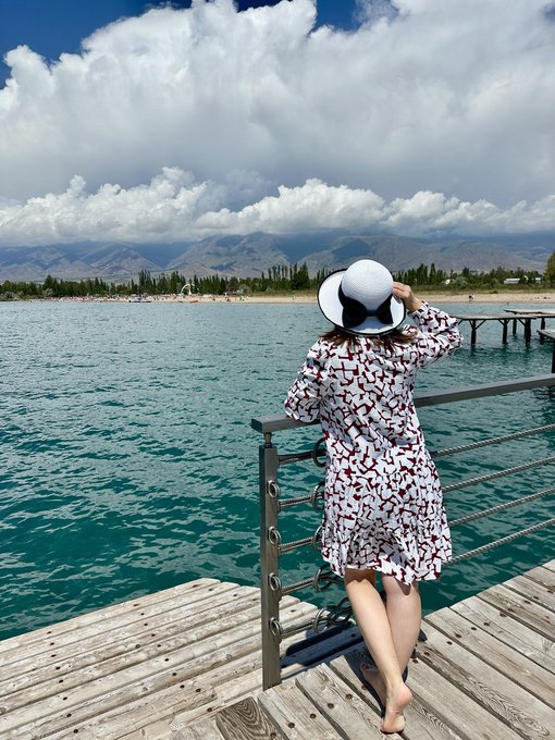 A woman wearing a white sun hat with a black bow and a patterned white dress stands on a wooden dock looking out over turquoise water toward mountains under a partly cloudy sky.