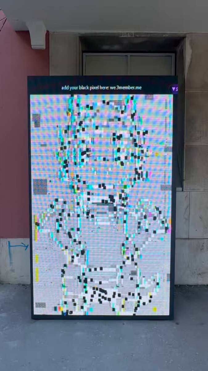 The image features a large, colorful digital art piece displayed outdoors. The art piece consists of small squares or rectangles that are colored green, blue, white, and black. These squares are arranged in various patterns and shapes, creating an abstract representation of a person's face. The text "Add your own place here where others have failed" is written above the artwork, suggesting that viewers can contribute to the creation of this piece by adding their own unique designs or ideas.