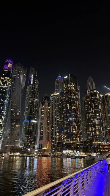 A nighttime cityscape view of illuminated skyscrapers along a waterfront with boats, featuring blue lighting on a railing in the foreground.