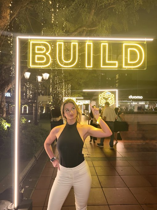 A woman poses confidently at night beneath a glowing yellow neon "BUILD" sign, with a "duomo" storefront and decorative tree canopies in the distance. The scene visually echoes the post's call to "build" at BNB, capturing an energetic, community-focused moment tied to the $BNB ecosystem.