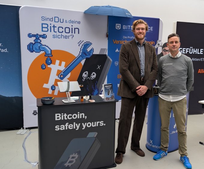 Two men standing next to a Bitcoin promotional display with the text "Sind DU & deine Bitcoin sicher?" and "Bitcoin, safely yours," featuring pixel art of a Bitcoin faucet and wrench, with BITSURANCE branding visible.