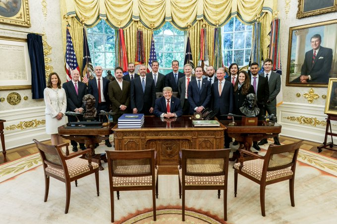A formal group photo in the Oval Office featuring a man seated at the Resolute Desk with approximately fifteen people standing behind him, with American flags and gold curtains in the background.
