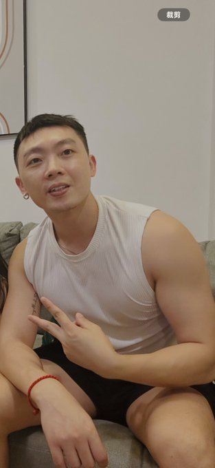 A short video screenshot (marked '裁剪') showing a muscular man in a white tank top making a peace sign while sitting on a couch with another person beside him.