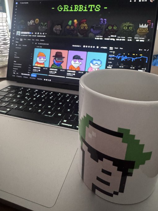 A white mug featuring a pixel art character design sits in front of an open laptop displaying the 'GRIBBITS' NFT collection page with cartoon character thumbnails.