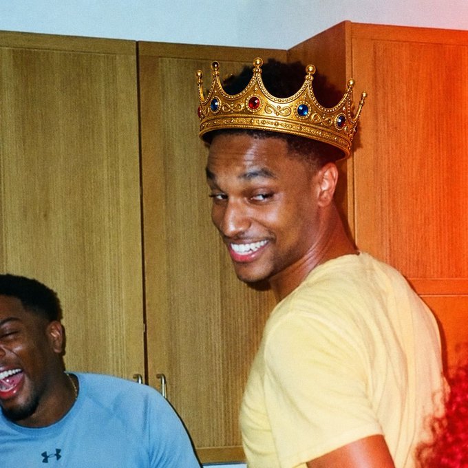 A smiling young man wearing a yellow t-shirt and a decorative gold crown with colorful jewels, standing in what appears to be a locker room with another person laughing beside him.