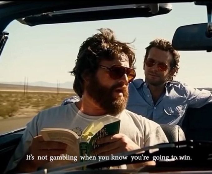 Two men ride in a convertible through a desert landscape, with the bearded driver reading 'The Richest Man in Babylon' while a subtitle reads, 'It's not gambling when you know you're going to win.'