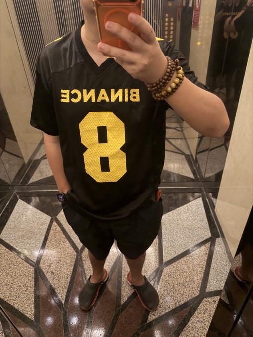 The image features a young man taking a selfie with his cell phone, wearing a black jersey with the number 8 printed on it. The background of the photo shows an elevator or escalator, suggesting that they are in a public place like an office building or shopping center. The caption to the image reads "GM 虚拟货币终究是黄粱一梦、庞氏骗局 @binancezh". This caption is likely related to a post on social media, where the young man has taken a photo of himself wearing his jersey and posted it online.