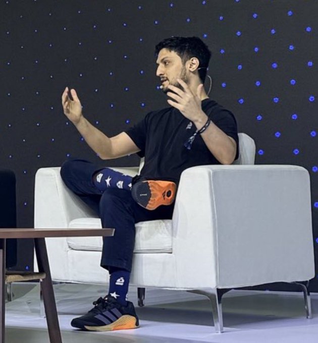 A man with a beard, wearing a black t-shirt, patterned socks, and orange-and-black sneakers, sits in a white armchair on stage, gesturing animatedly with raised hands against a dark backdrop dotted with blue lights.

The image shows the man mid-discussion on a panel or interview stage, illustrating the tweet's message about @Backpack's vision to list a stock on Solana for continuous, 24-hour trading beyond traditional Nasdaq sessions.