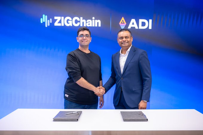 Two men shaking hands in front of a blue backdrop featuring the ZIGChain and ADI logos, with folders on a white table in the foreground.
