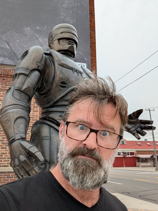 A bearded man with glasses takes a selfie in front of a large metallic robot statue, appearing surprised.