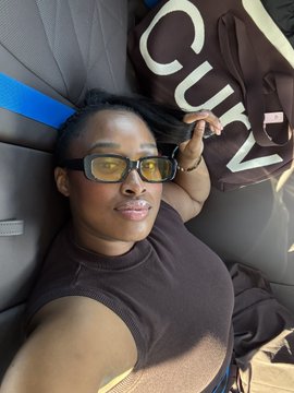 A selfie of a woman wearing yellow-tinted glasses and a dark tank top, reclining in a chair next to a black bag with 'COVR' visible on it.
