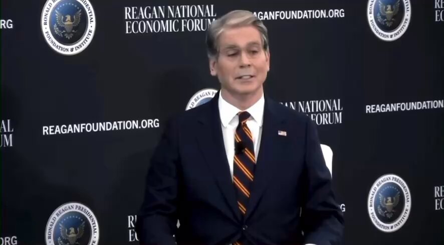 The image features a man dressed in a suit and tie standing against a black background with white text. The man appears to be speaking or giving a speech, as indicated by his body language and facial expression. The backdrop includes logos for organizations such as the Reagan Foundation and the Regan National Economic Forum, suggesting that this event may have been organized by these groups.