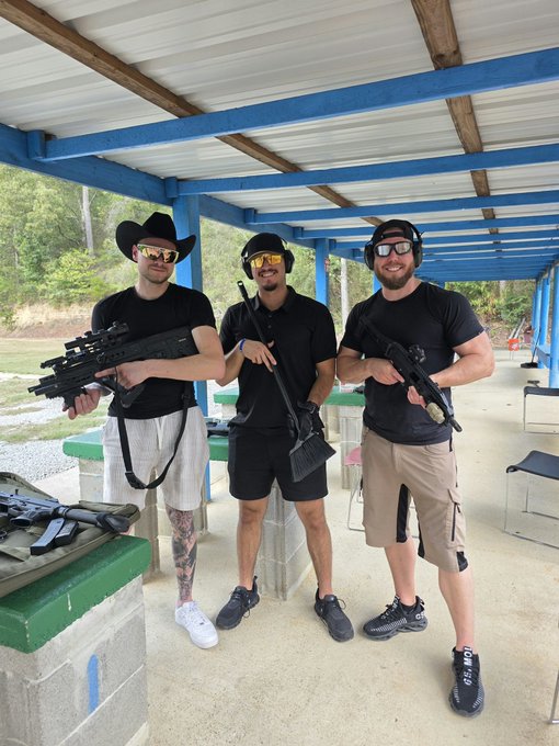 Three men pose at an outdoor shooting range wearing protective earmuffs and yellow safety glasses, each holding a different firearm.
