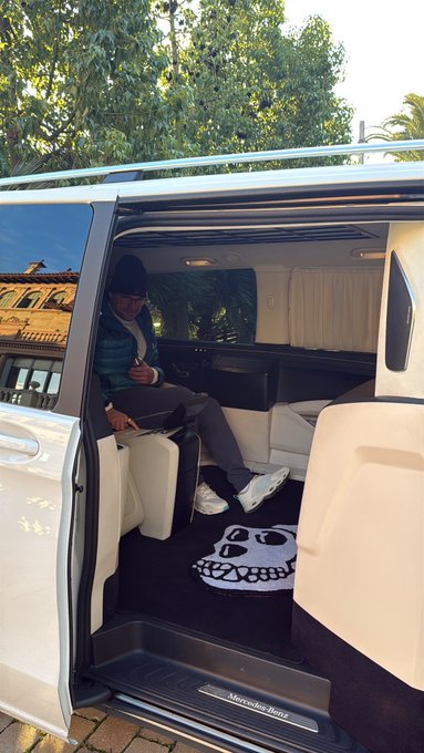 A man sitting inside a white Mercedes-Benz van with a custom skull-themed floor mat, with the sliding door open revealing a luxurious cream interior.