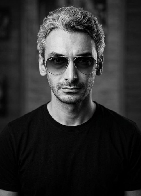 A black-and-white portrait of a stylish young man with wavy hair and aviator sunglasses, wearing a black t-shirt and looking directly at the camera.