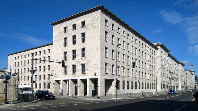 A large, modern, multi-story stone building with numerous rectangular windows stands at a street corner under a clear blue sky.