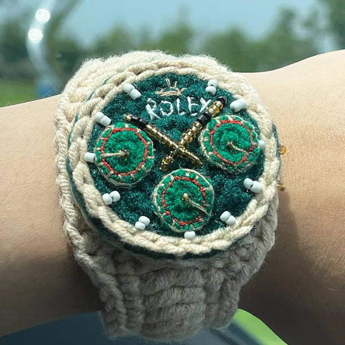 A crocheted yarn replica of a Rolex watch on a person's wrist, featuring green 'dials' and the word 'ROLEX' embroidered on the face.