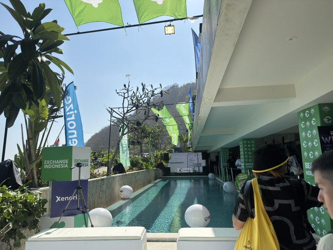 Event staff stand poolside at a Tokocrypto Horizon venue in Indonesia, with banners promoting "Exchange Indonesia" and "Xeni" alongside green Tokocrypto flags, suggesting a sunny but tolerable crypto industry gathering.