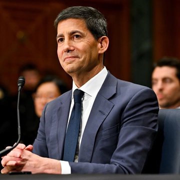 A man in a dark suit and tie with short graying hair sitting at what appears to be a congressional hearing.
