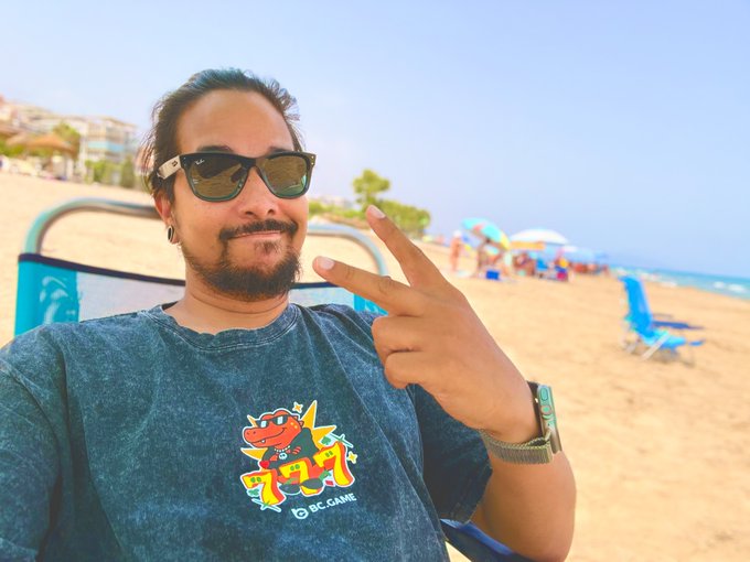 A bearded man in sunglasses poses with a peace sign at the beach, wearing a "BC.GAME" t-shirt and flashing a wristwatch, capturing a relaxed "GM" (good morning) vibe with palm trees and umbrellas in the background.