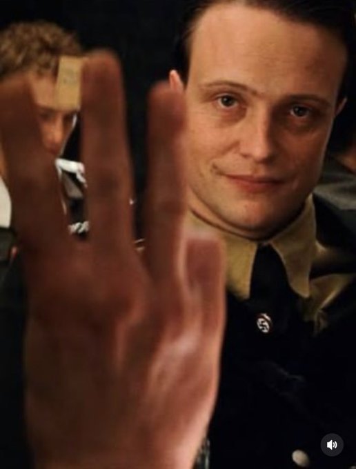 A scene from a film showing a man in a dark uniform with a Nazi pin on his collar smiling and extending three fingers toward the camera.