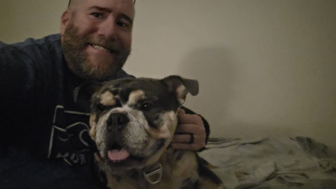 A smiling bearded man poses for a selfie with a brindle and white bulldog whose tongue is hanging out.