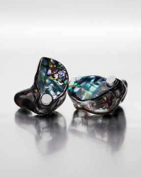 Two in-ear monitors with iridescent abalone faceplates rest on a reflective white surface, their swirled blue, green, and purple hues perfectly illustrating the post's claim that each Ripple Abalone faceplate is uniquely one-of-a-kind.