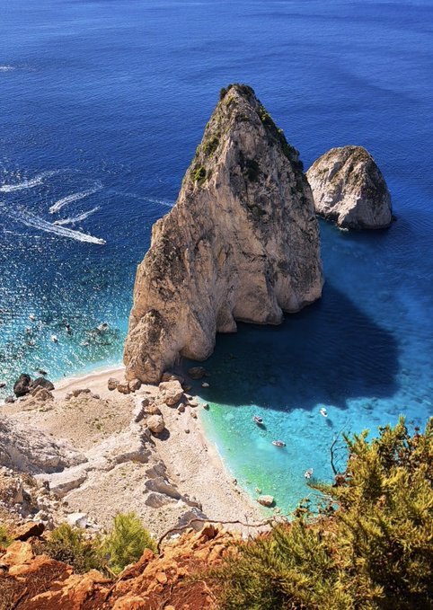 A towering sea stack rises dramatically from crystal-clear turquoise waters along a rocky coastline, with a few boats anchored in the cove below. The image is paired with a caption declaring "It's definitely a bull market," suggesting the majestic, upward-reaching rock formation symbolizes bullish financial momentum and rising strength.
