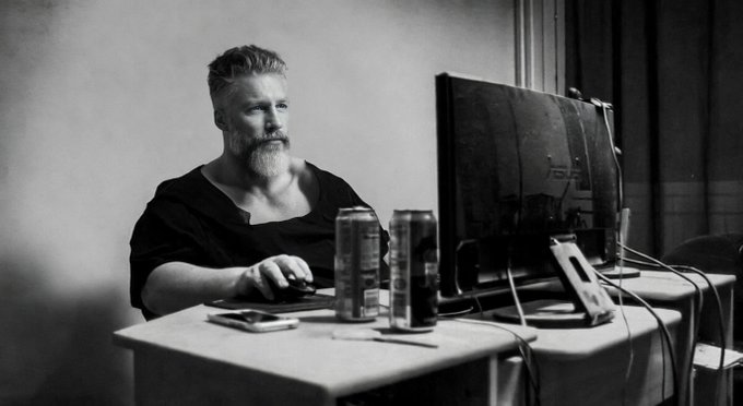 A black and white photo shows a bearded man sitting at a desk using a computer, with energy drink cans placed next to the monitor.