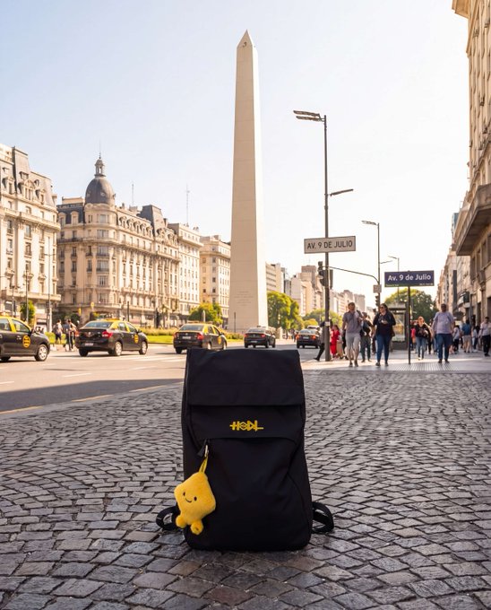 A black backpack with a yellow plush charm rests on a cobblestone street in Buenos Aires, framed by the iconic Obelisco monument and street signs reading "Av. 9 de Julio," serving as visual proof of travel.