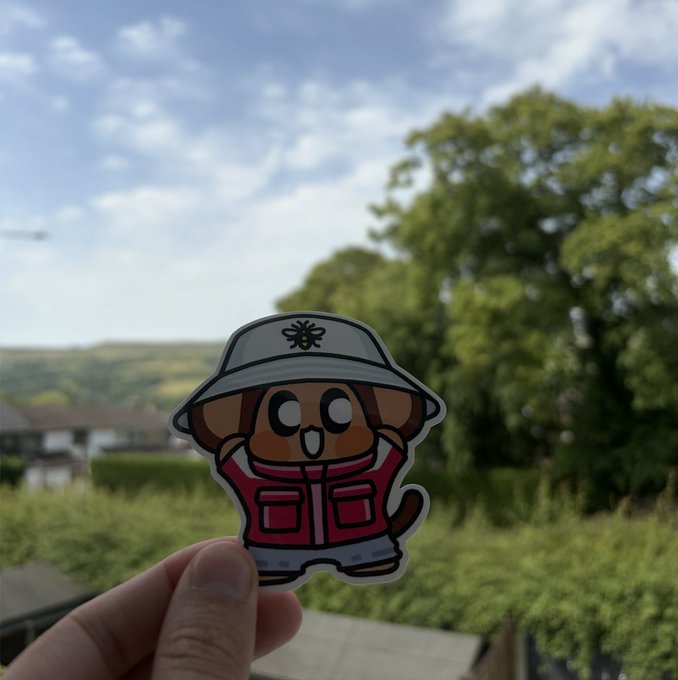 The image features a person holding up a sticker featuring a cartoon dog wearing a hat and a red jacket with pockets. The background shows trees, suggesting that they are outdoors. The caption of the image reads "Looking forward to the @ChimpersHQ x @FugzOfficial meetup in Manchester this Saturday! If you’re in the UK, come hang out with us!".