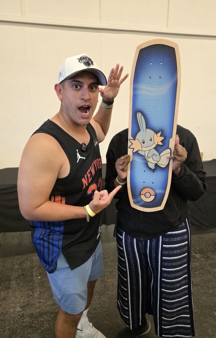 An excited man in a black Knicks jersey and white cap points at a Pokémon-themed skateboard deck featuring Mudkip held by another person.