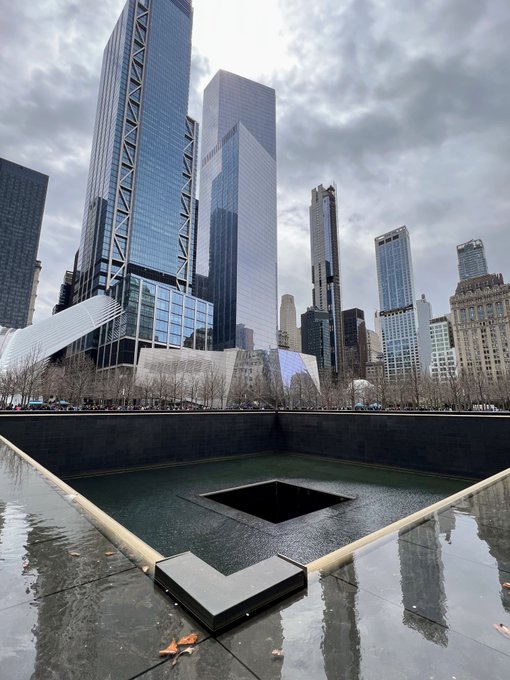 A square reflecting pool at the 9/11 Memorial in New York City sits at the center of a void, its dark water cascading into an abyss framed by towering skyscrapers under an overcast sky. The image visually embodies the post's description of "an unfillable void," capturing the haunting emptiness and profound sense of loss at Ground Zero.